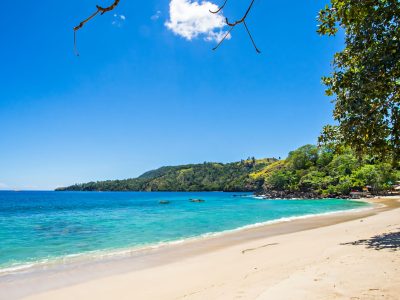 Panorama aerial Pantai Pulisan di Likupang dengan perairan biru toska, pasir putih, dan perbukitan hijau yang mengelilingi teluk.